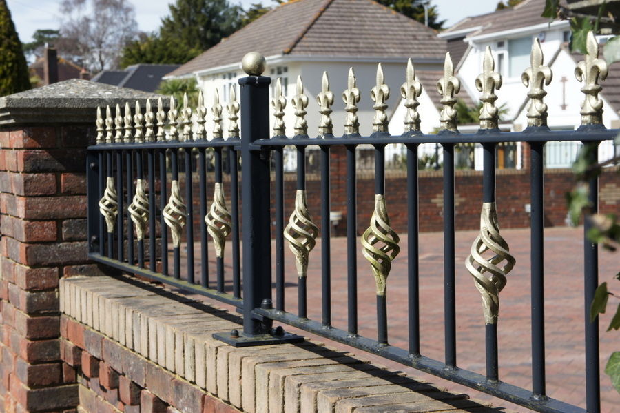 Black railings with gold details on a brick wall beside a block-paved drive