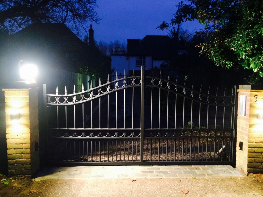 Black metal double driveway gates with a curved top and rings, lit by pier lights at dusk